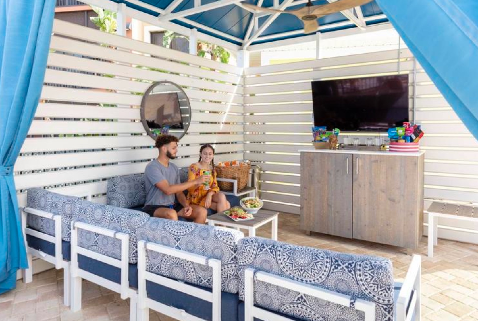 A couple enjoying drinks, sitting on the couch, in their poolside cabana